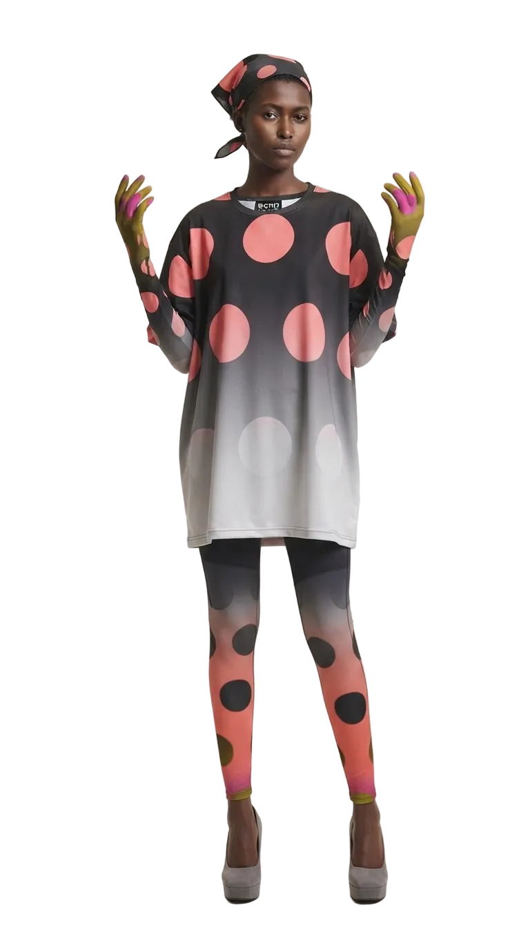 A model wearing a black and pink polka dot tunic top with a gray gradient hem, matching gradient leggings, and a black headscarf, standing against a light beige studio background.
