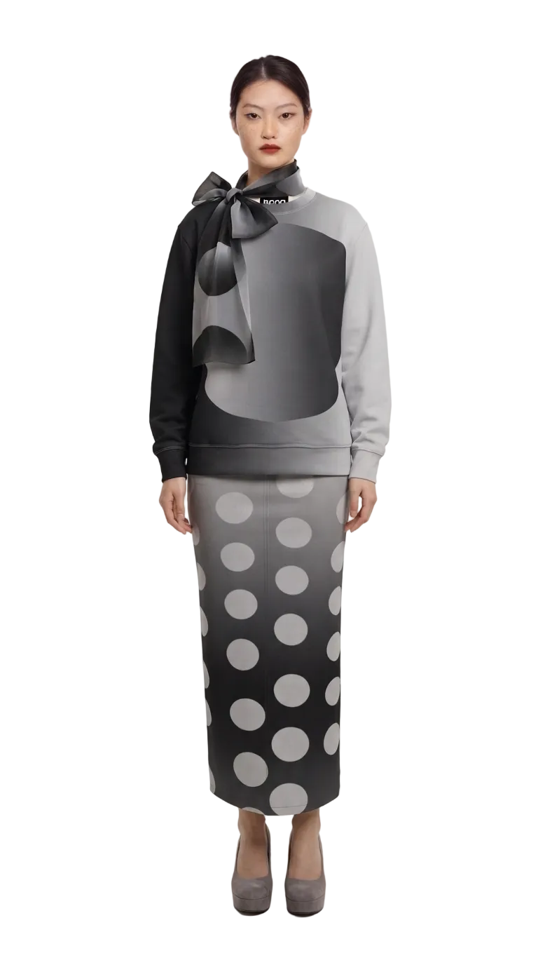 A model stands against a neutral studio backdrop wearing a matching gray and black outfit with a large bow at the collar and a skirt featuring a large polka dot pattern.